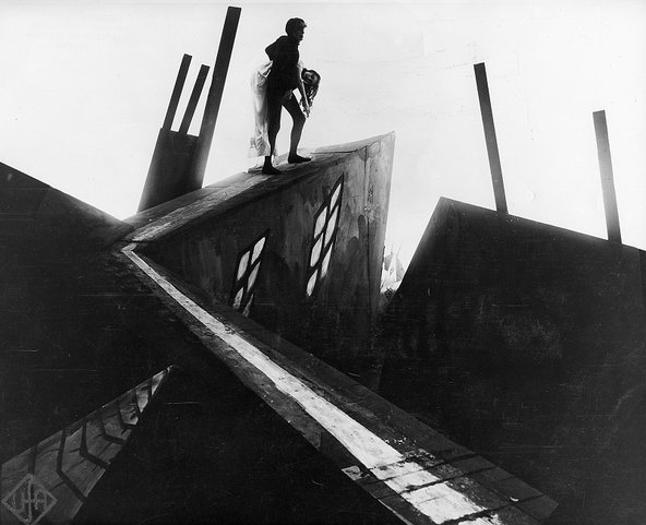 A haunting black-and-white film still showing outlines of a male figure, Dr Caligari, carrying an unconscious woman up the side of a building against a bright horizon. The elements of architecture and the angles are distorted in the image.