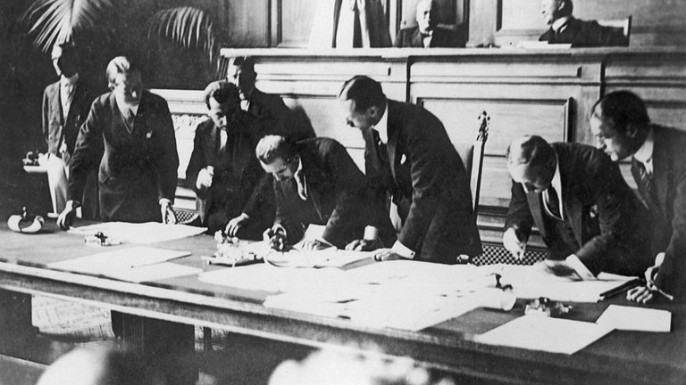 Black and white photo of the signing of the Treaty Of Lausanne In 1923 Eight men in suits are leaning over a table on which documents are laid out. .