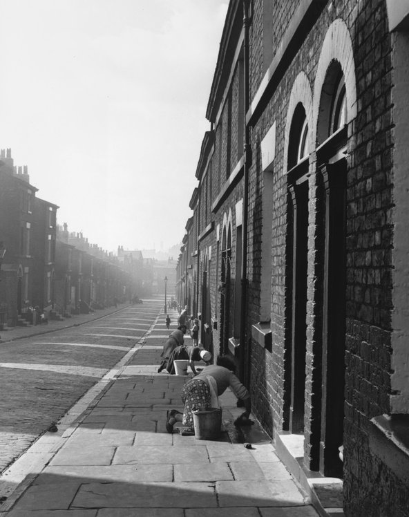 Black-and-white image of a long empty street with houses stretching into the horizon. In the shadows of the buildings, a row of women are scrubbing the doorsteps of their homes.